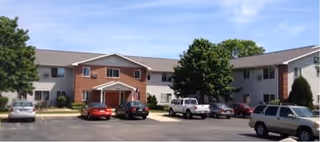 Exterior view of a two-story apartment building with a parking lot in front. Several cars are parked, and there are trees and shrubs around the building under a clear blue sky.