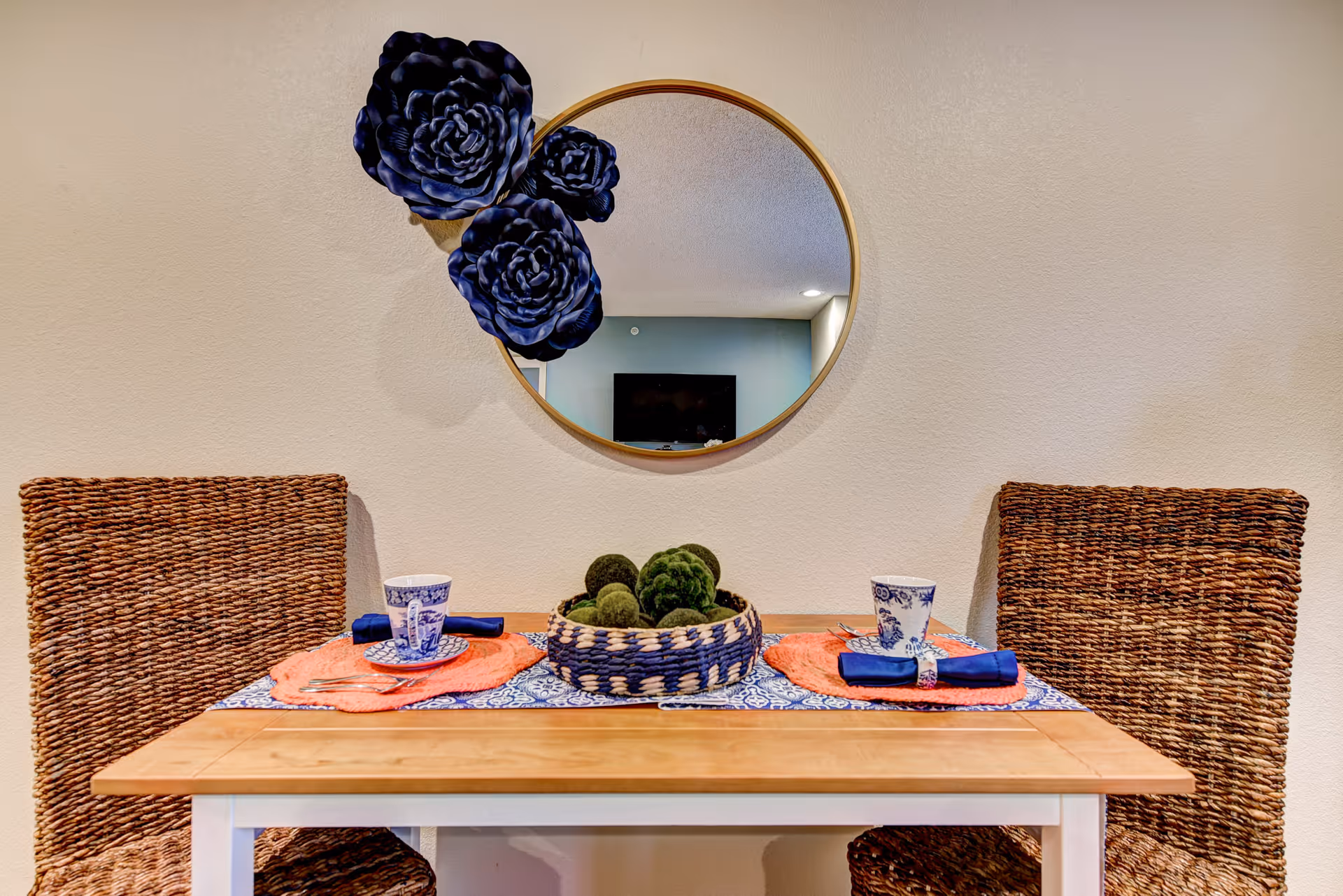 A dining table with wicker chairs, moss centerpiece, and round mirror on beige wall.