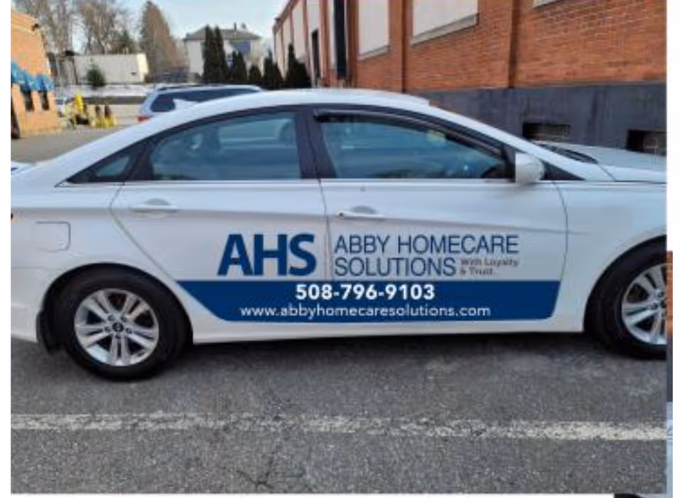 White car with AHS Abby Homecare Solutions branding parked in a lot beside a brick building.