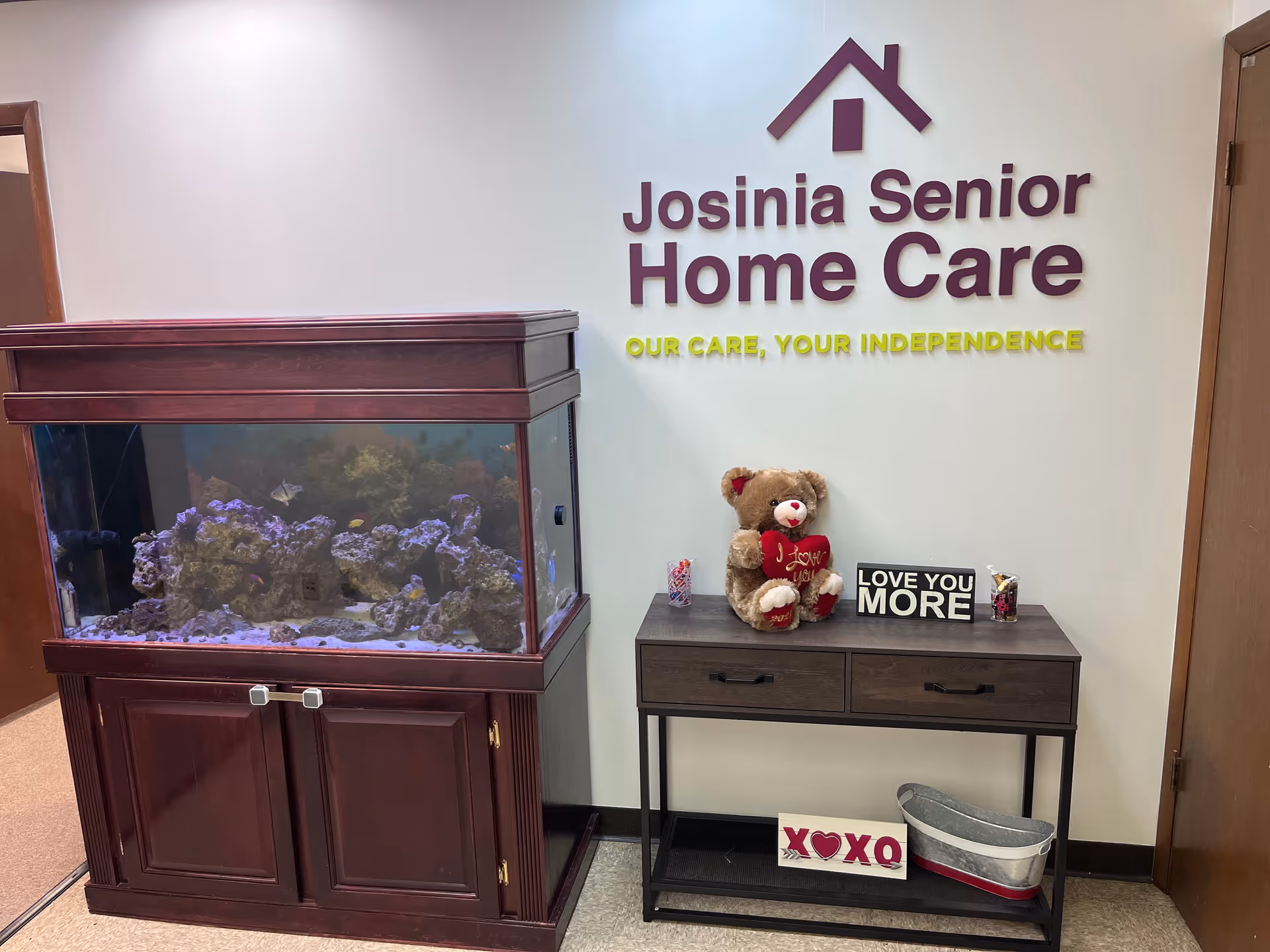 Interior lobby area with Josinia Senior Home Care sign, aquarium, teddy bear, and decorative table.