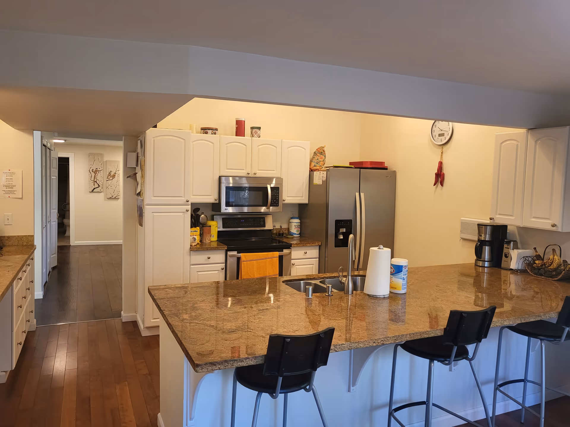Open kitchen with a large granite island and three bar stools, white cabinets, stainless steel refrigerator and microwave, and a hallway in the background.