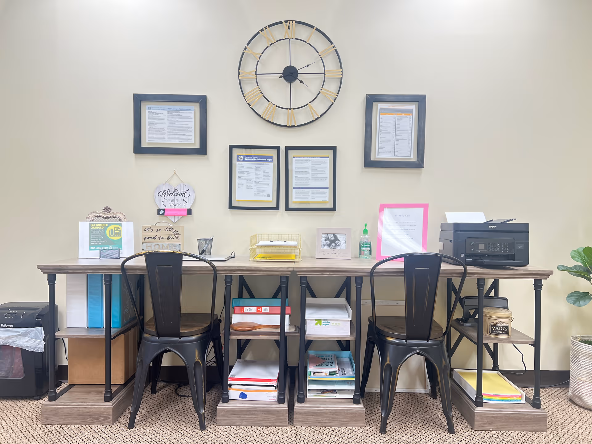 A long desk against a wall with framed documents, a large clock, and three metal chairs.