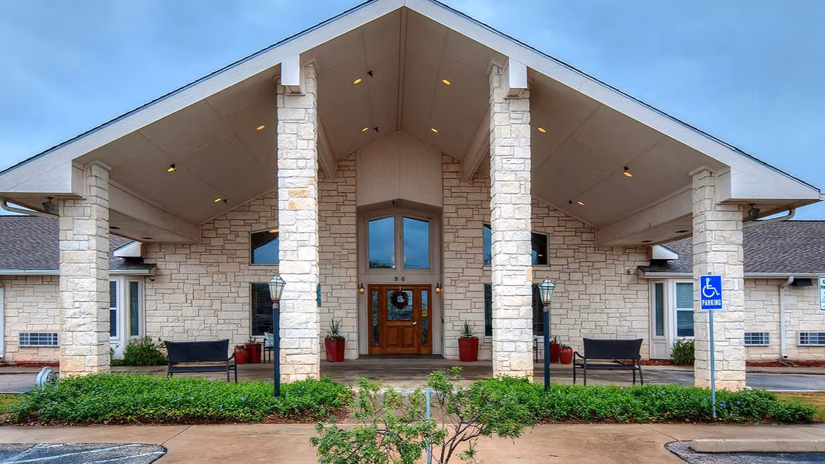 Stone-faced building entrance with a large covered portico, tall pillars, double wooden doors, benches and landscaping.