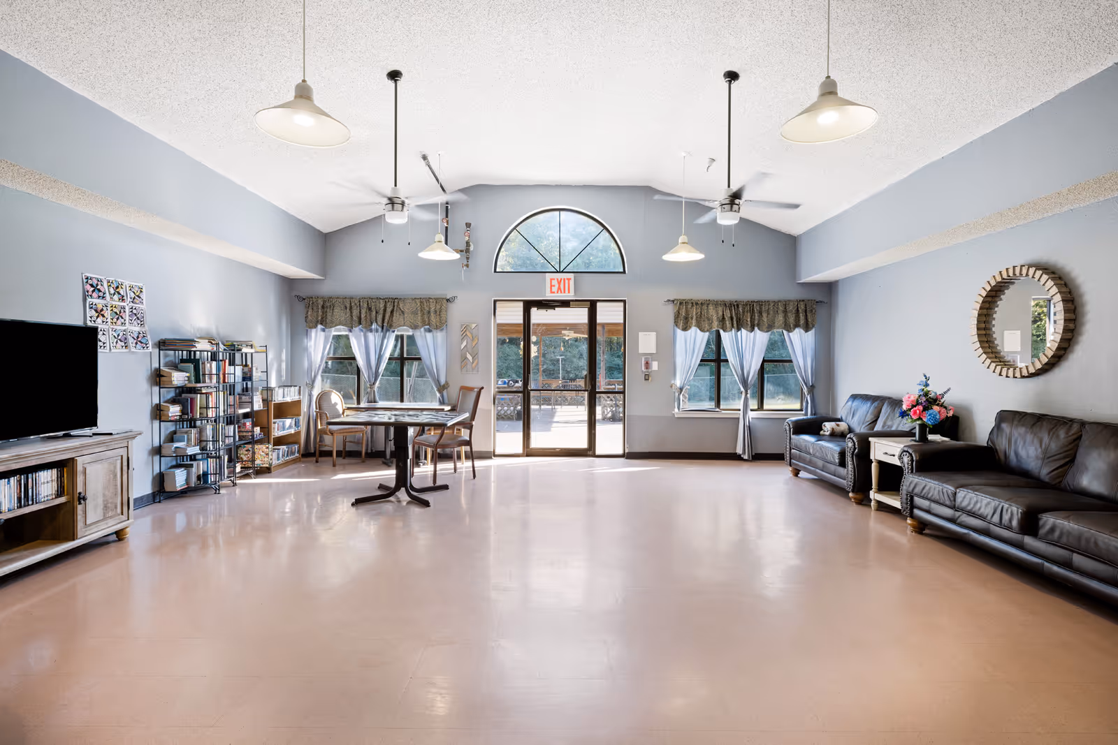 Bright communal lounge with sofas, a table, bookshelves and double exit doors beneath an arched window.