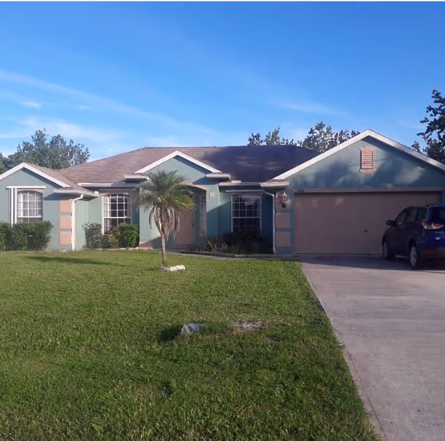 Single-story teal house with an attached garage, driveway with a parked car, a front lawn and small palm tree under a clear blue sky.