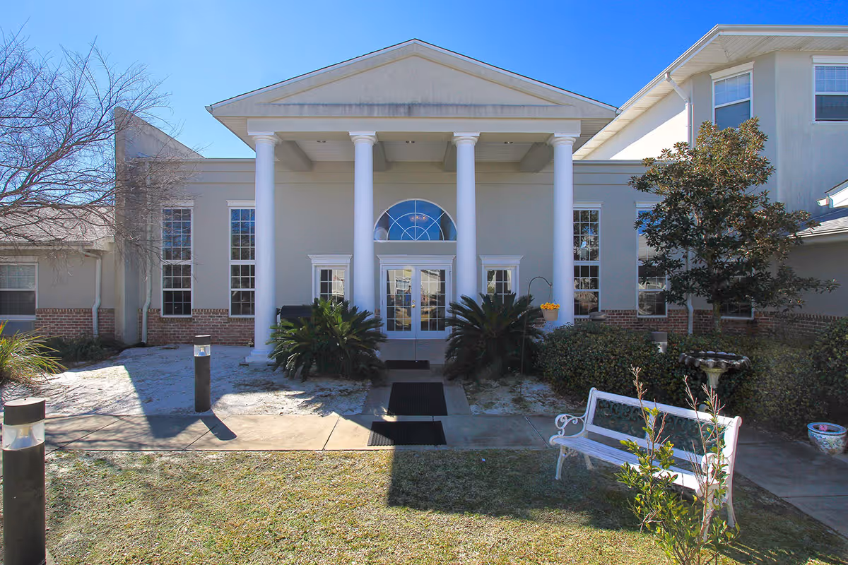 Front exterior view of Gulf Breeze Courtyard Assisted Living building with white columns, large windows, a bench, and landscaping including bushes and a tree under a clear blue sky.