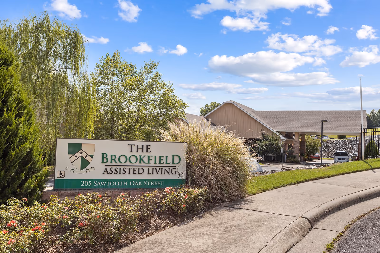 Exterior view of The Brookfield Assisted Living facility sign surrounded by landscaping with bushes and flowers, a paved sidewalk, and the building entrance visible in the background under a blue sky with scattered clouds.