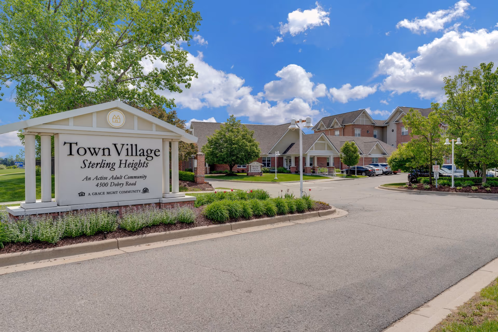 Entrance sign for Town Village Sterling Heights in front of the community buildings and driveway under a blue sky.