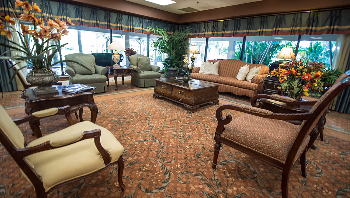 Cozy senior living lounge with sofas, chairs, plants, and a central coffee table.