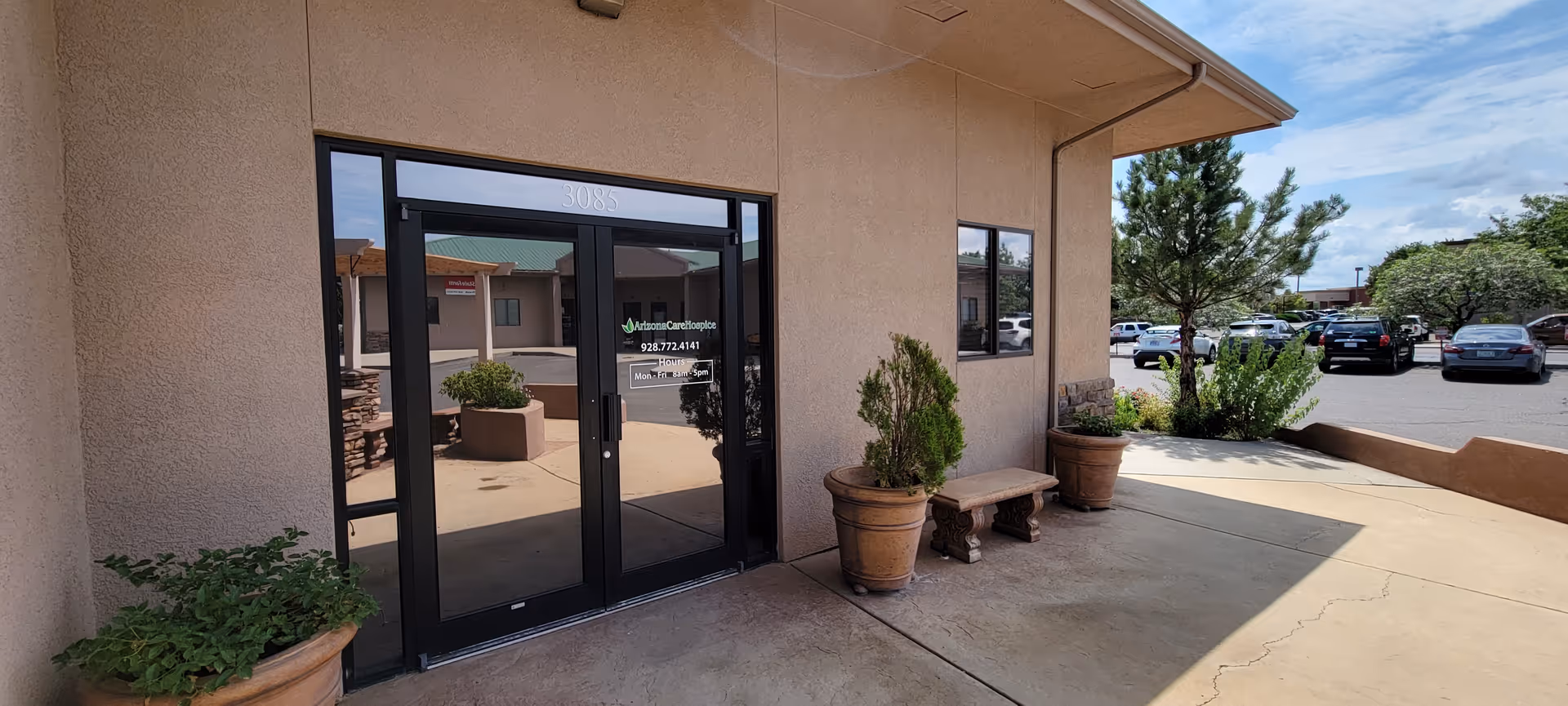 Exterior view of a hospice facility entrance with glass doors, potted plants, and a parking lot.