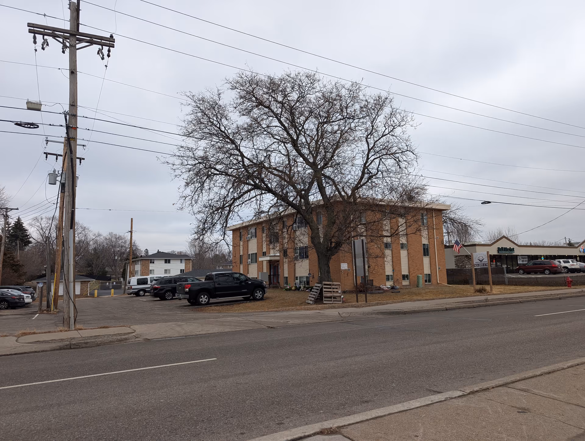 Exterior view of a three-story brick building with several windows, a large leafless tree in front, and a parking area with multiple vehicles. The sky is overcast, and there are power lines and a utility pole visible along the street in front of the building.