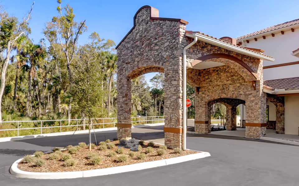 Stone-arched porte-cochere entrance of a senior living building with a landscaped traffic island and trees beyond.