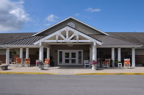 Front exterior view of a single-story building with a covered entrance supported by white columns. There are several rocking chairs and flower pots arranged on the porch area under a clear blue sky with some clouds.