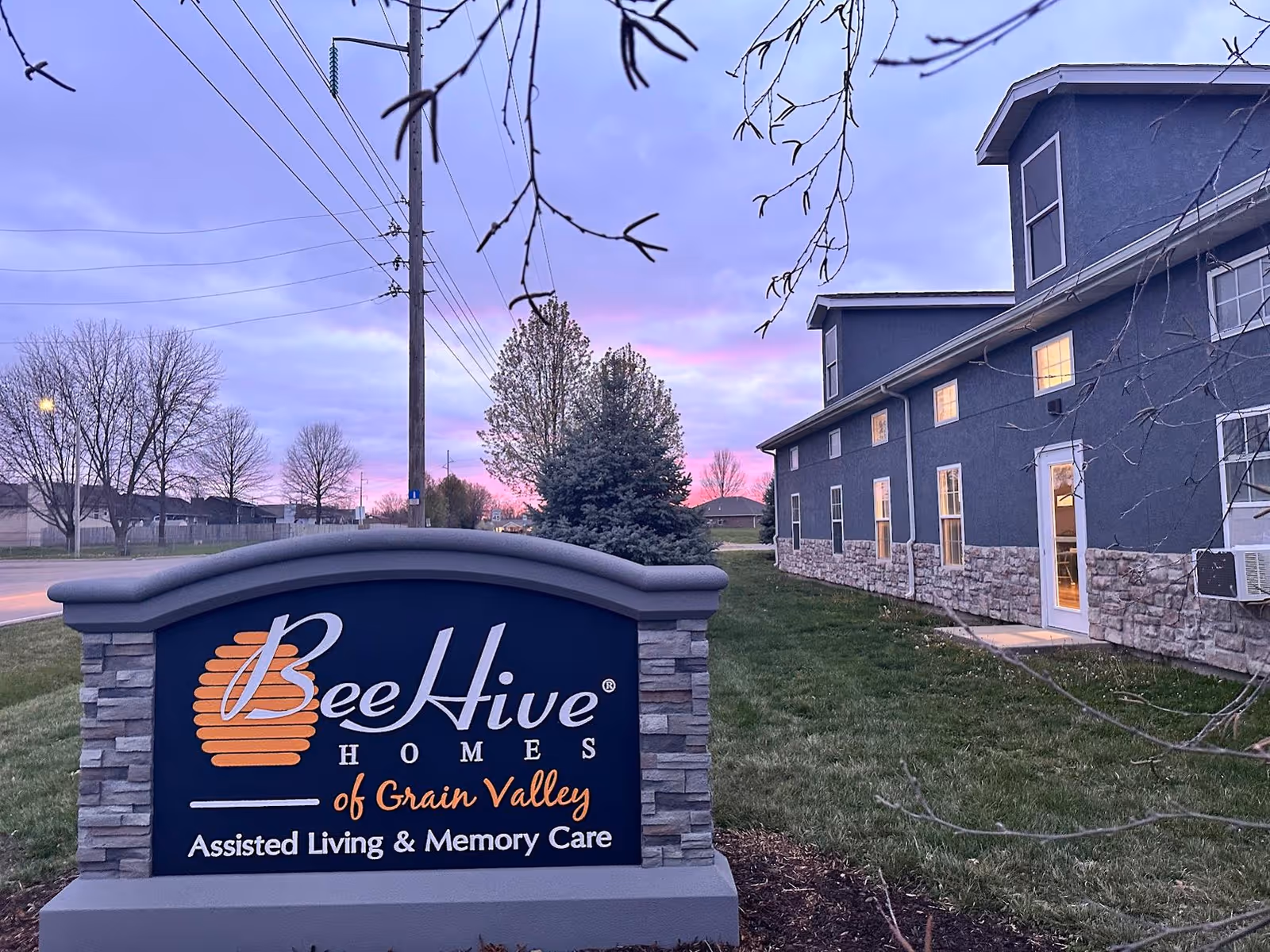 Exterior view of BeeHive Homes of Grain Valley assisted living and memory care facility at dusk, showing a stone and gray building with lit windows and a large sign in the foreground with the facility name and services.