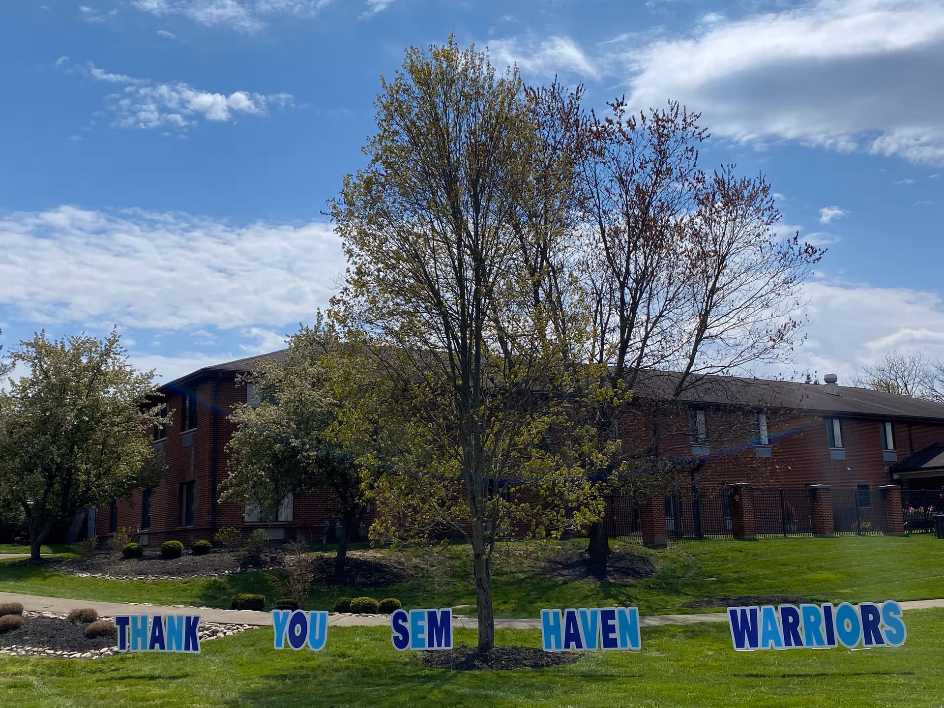 Brick senior living building with trees and a Thank You SEM Haven Warriors sign.