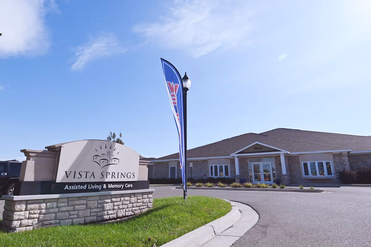 Exterior view of Vista Springs Assisted Living & Memory Care facility showing the main building with a stone facade and a sign in front on a sunny day with a blue sky.