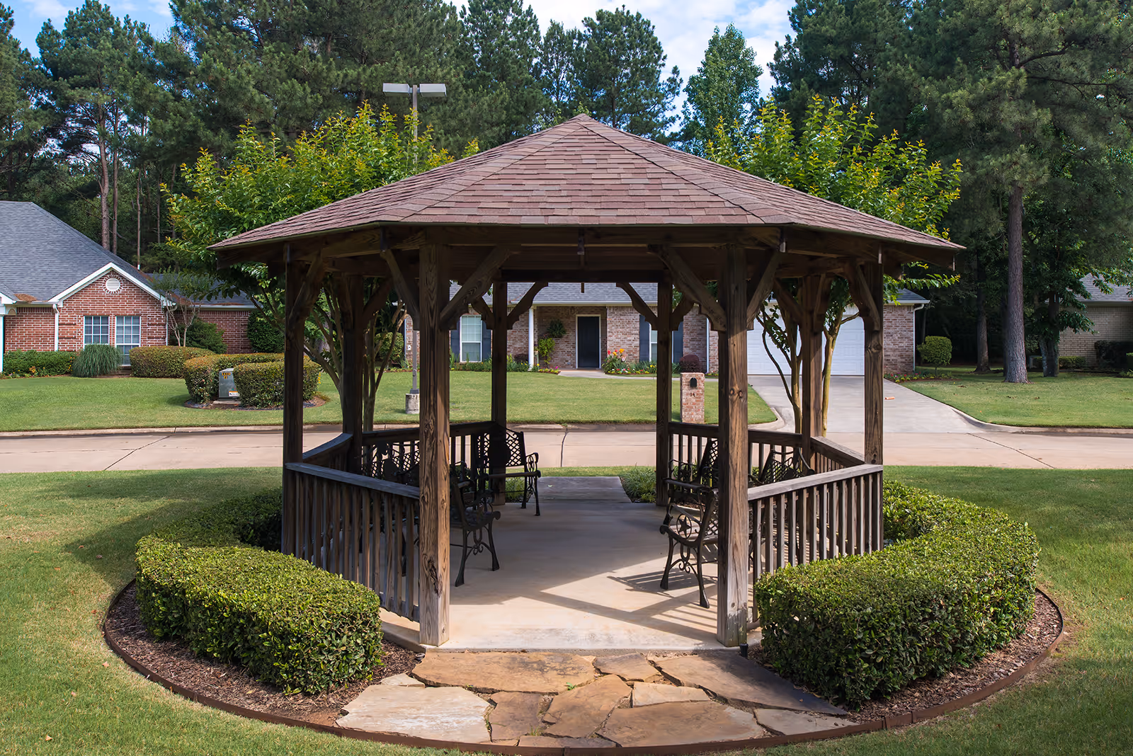 A wooden gazebo with a shingled roof situated in a well-maintained grassy area with trimmed bushes around it. There are metal benches inside the gazebo. In the background, there are residential houses, trees, and a paved road.