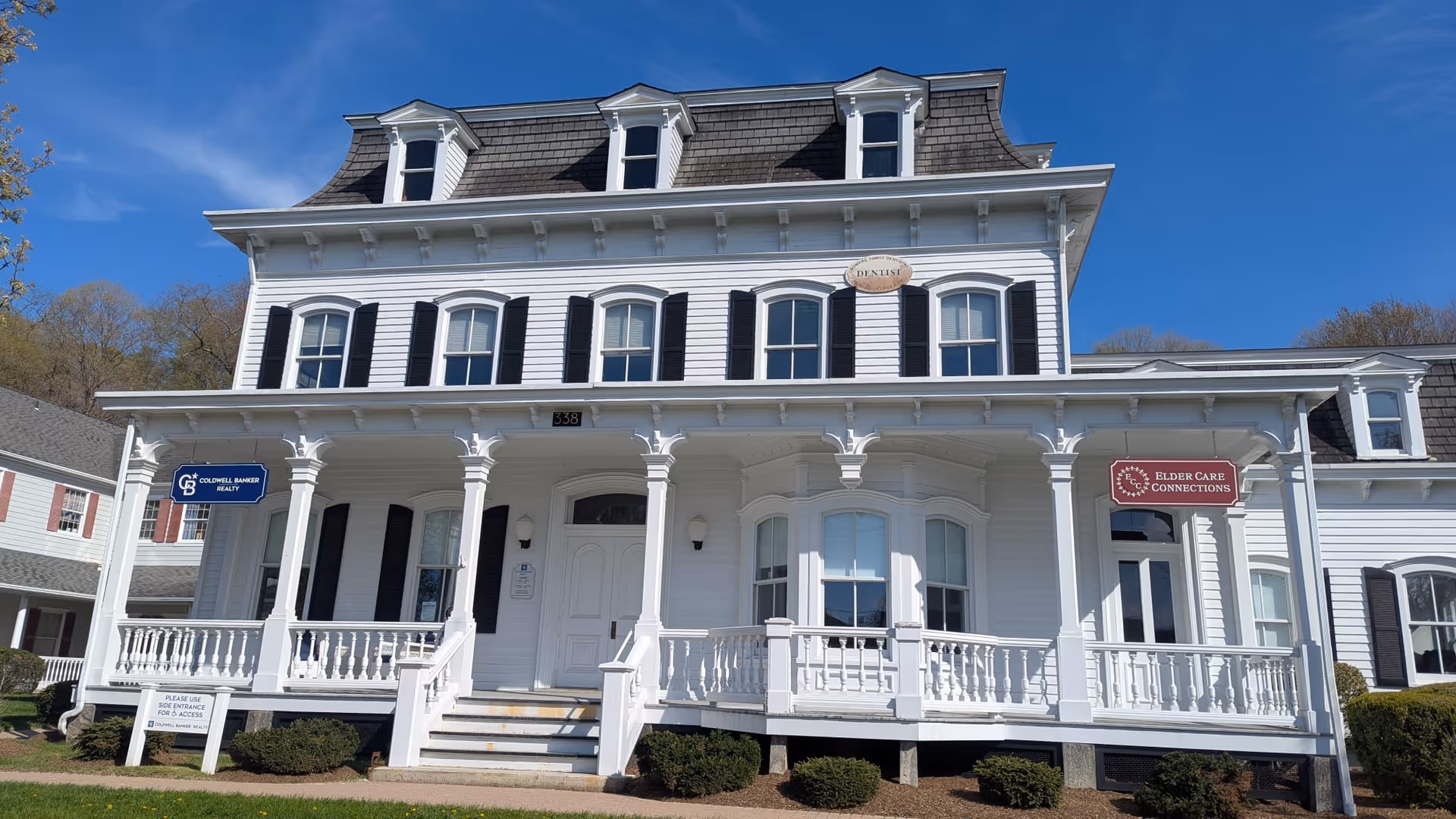 White, two-story Victorian-style building with a wraparound porch and dormer windows under blue sky.