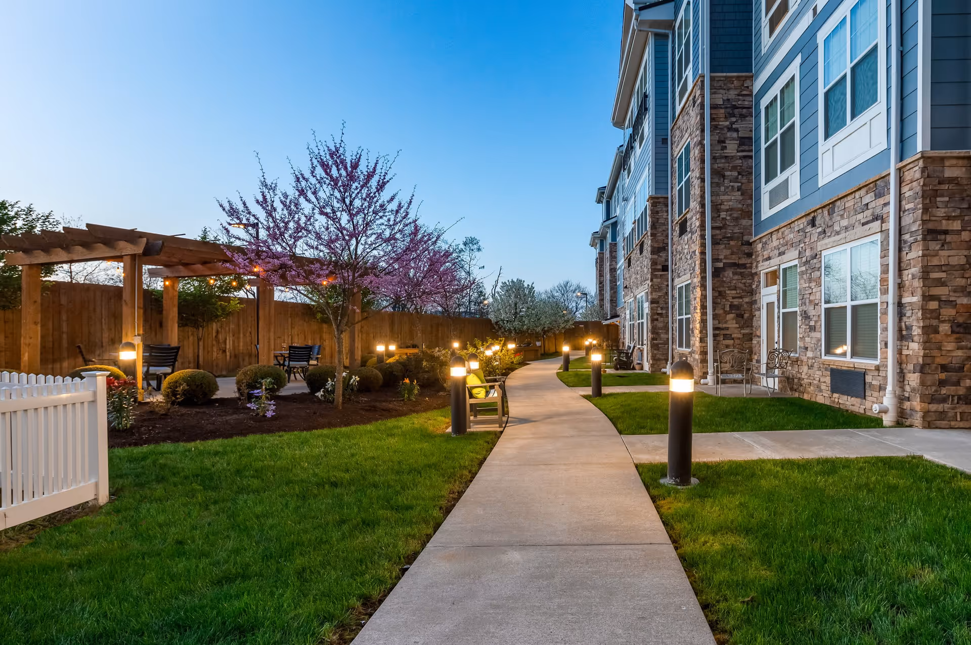 A landscaped courtyard walkway beside a multi-story stone-and-siding senior living building with benches, a pergola, and illuminated path lights.