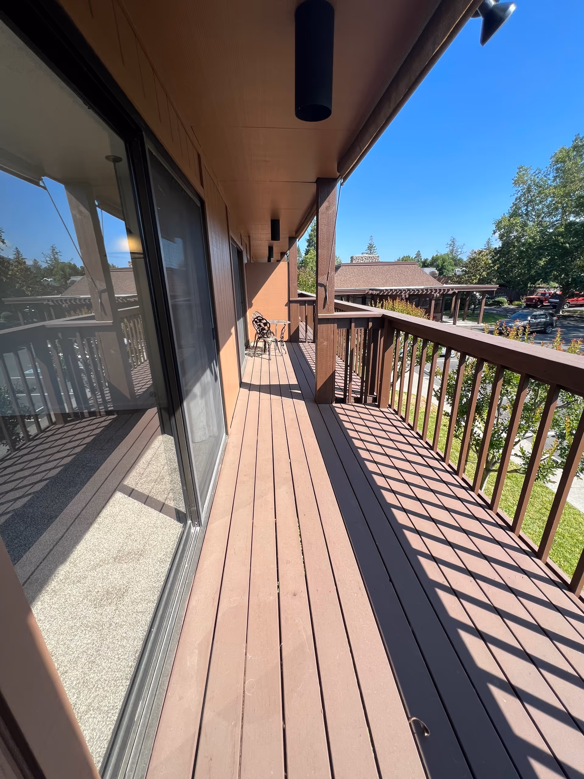 A long wooden balcony with brown railings and floorboards, attached to a building. There are two black metal chairs at the far end of the balcony. The balcony overlooks a parking area with cars and trees under a clear blue sky.