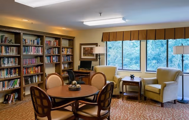 A cozy library and sitting area in Deer Park Assisted Living featuring wooden bookshelves filled with books, a round wooden table with four chairs, two beige armchairs near large windows with a view of green trees, a small wooden side table, a floor lamp, and a desk with a computer.