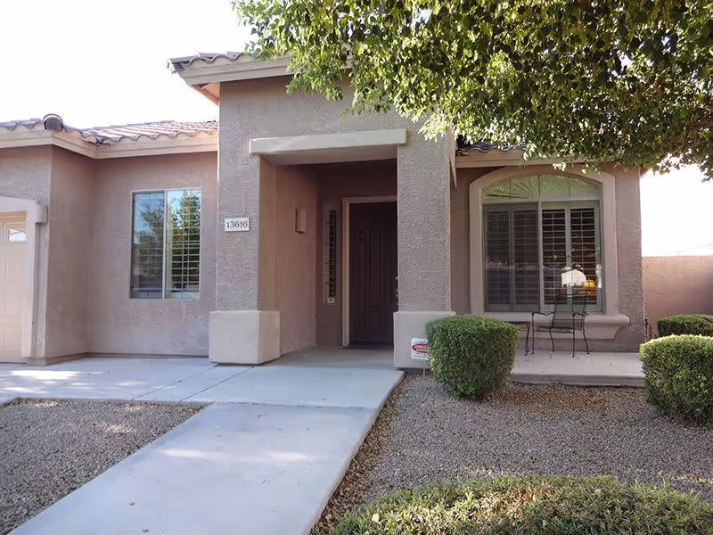 Front exterior view of a single-story residential building with a covered entrance, two windows with shutters, a small patio area with a table and two chairs, and landscaping including bushes and a tree providing shade.
