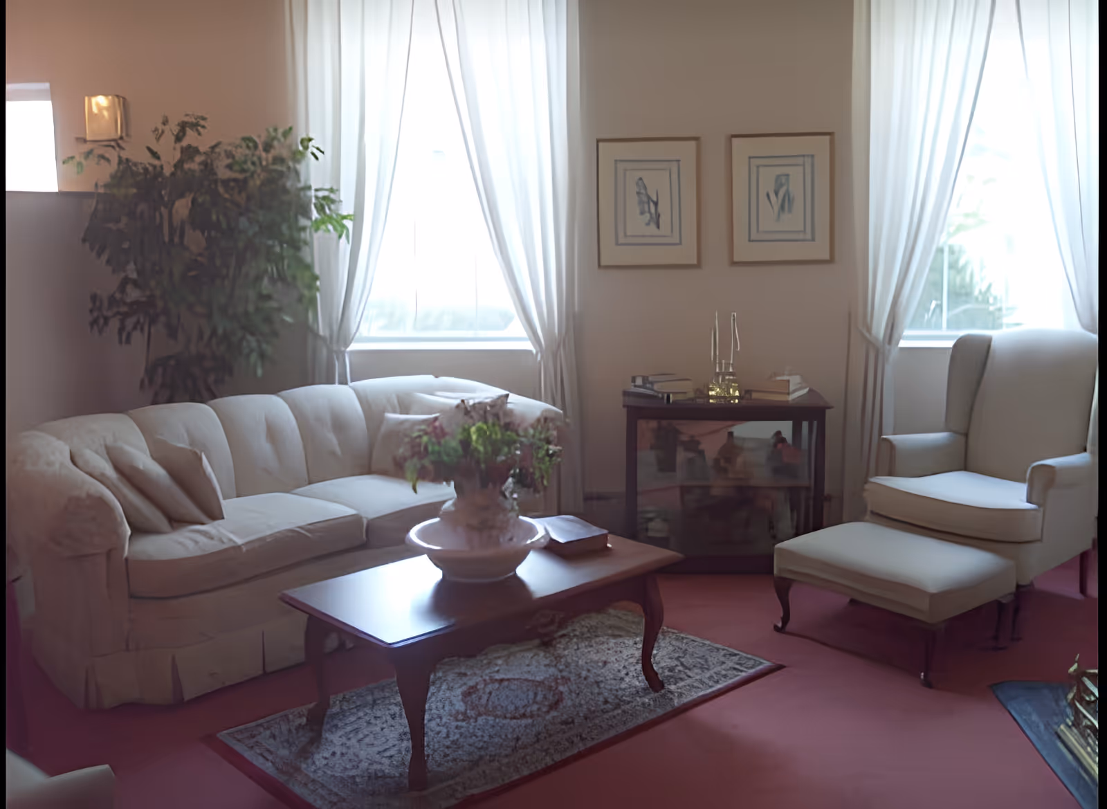 Sunlit traditional living room with a tufted sofa, armchair and ottoman, coffee table with a floral centerpiece, and curtained windows.