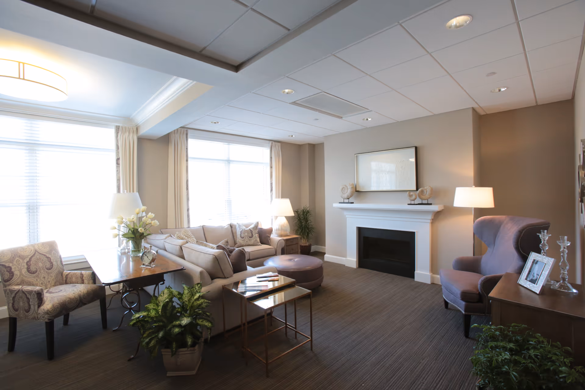 Bright hospice living room with sofa, chairs, fireplace, lamps, and large windows.