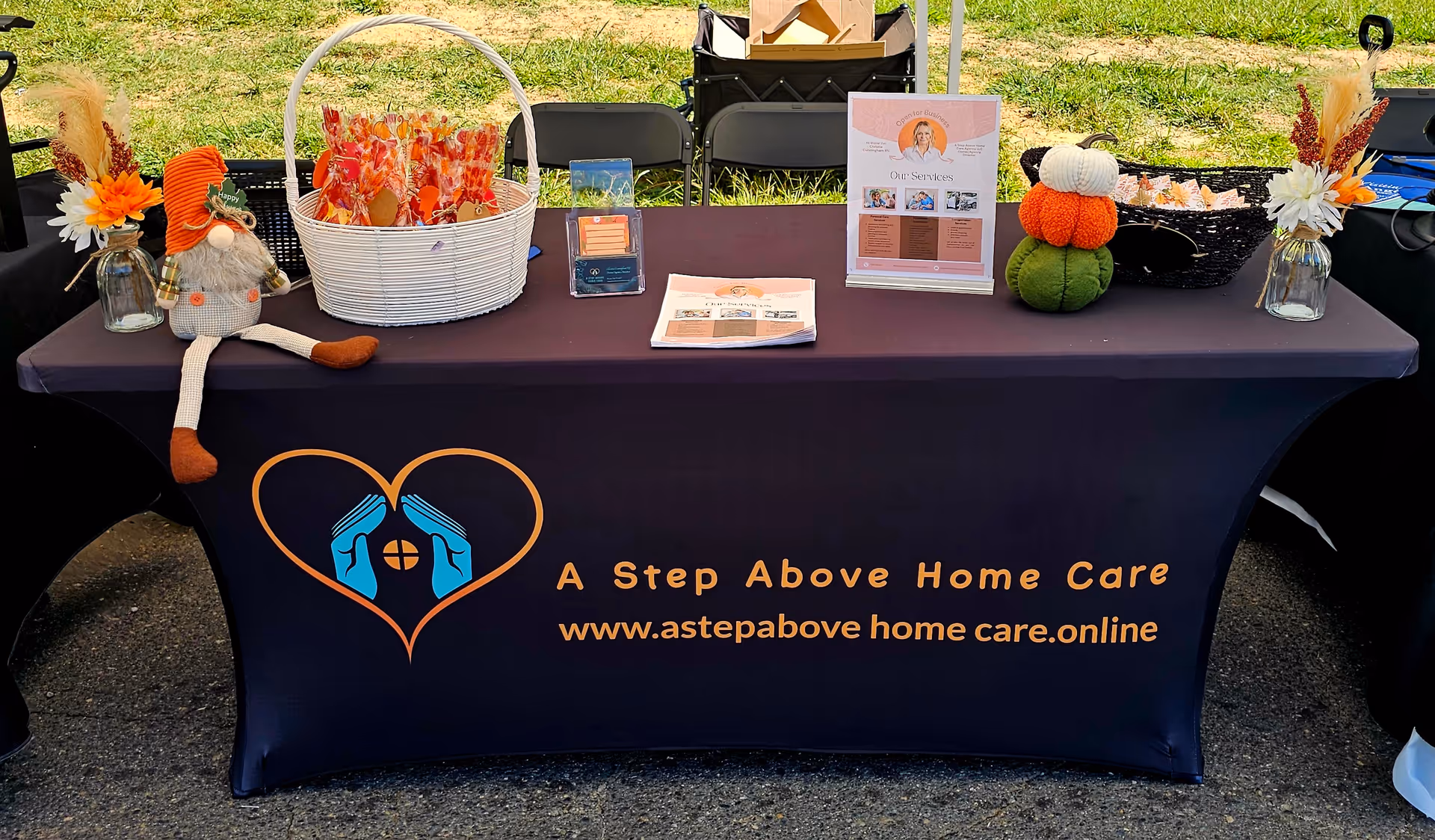 Outdoor booth table draped in navy with A Step Above Home Care logo and decorative items.