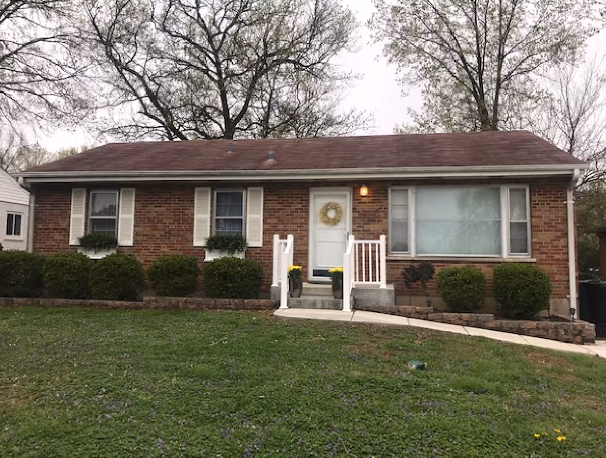 Brick one-story senior living home with white door, wreath, stairs, and trimmed shrubs.