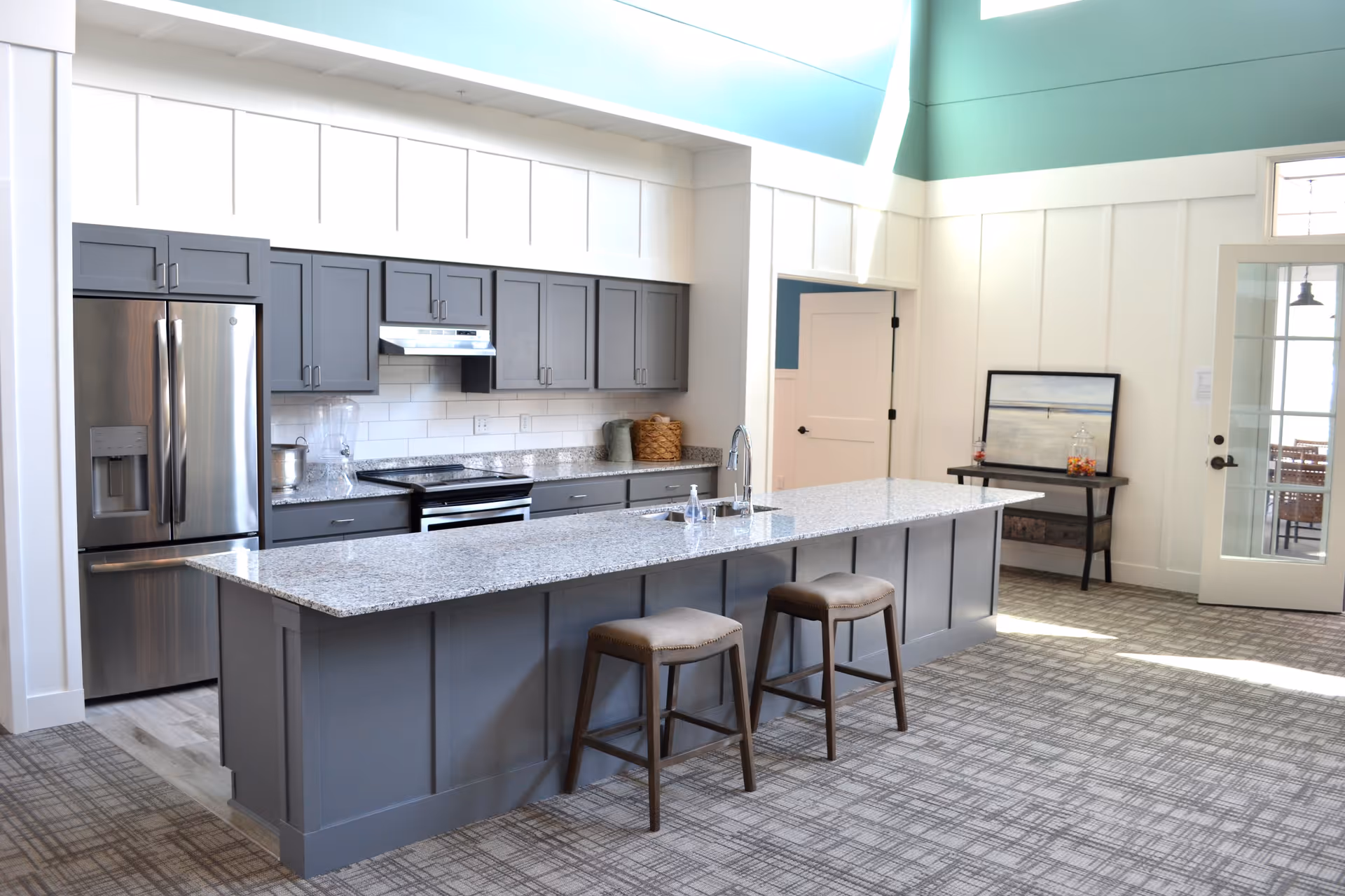 Modern kitchen area with gray cabinets, a large granite island with two stools, stainless steel refrigerator and oven, and a small table with a framed picture and candy jars near a glass door.