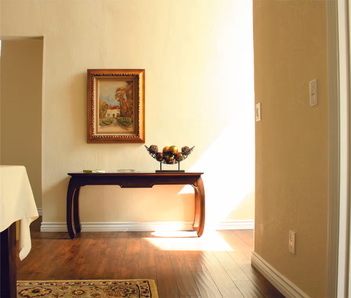 Sunlit interior hallway with a wooden console table topped with a fruit bowl and a framed painting on the wall.