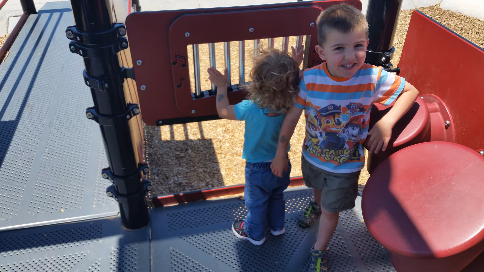 Two young children stand on a colorful outdoor playground structure, smiling at the camera.