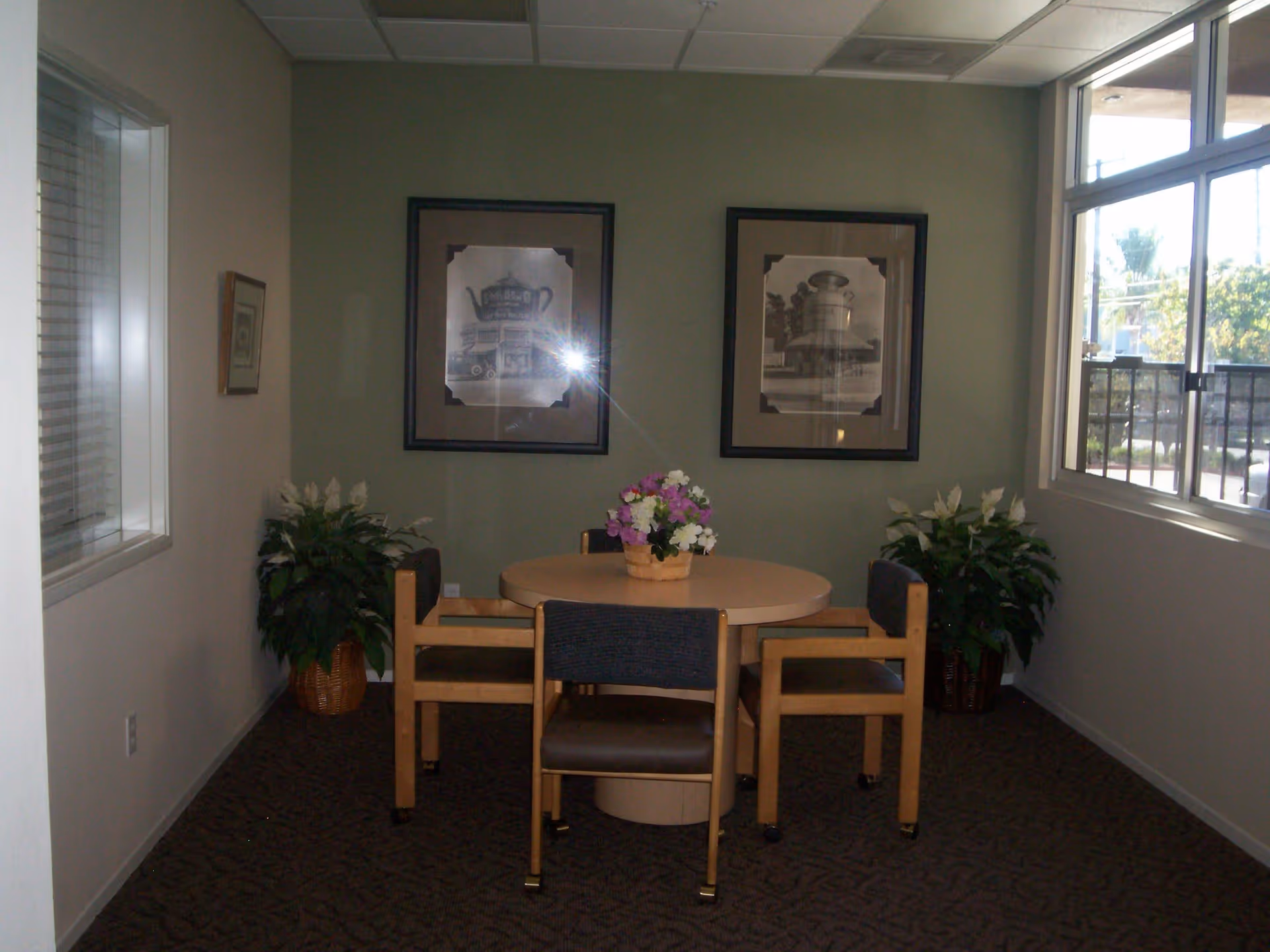 Small dining nook with a round table and four chairs, framed artwork on the wall, potted plants, and a window.