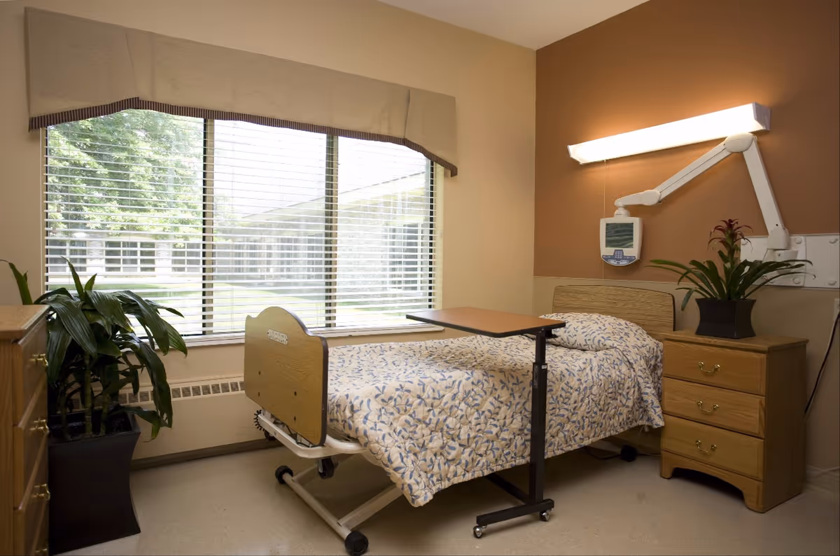 A healthcare facility room with a single hospital bed covered in a floral patterned blanket, an adjustable overbed table, a wooden nightstand with a potted plant, a large window with blinds and a beige valance, and a wall-mounted medical device with an adjustable arm light.