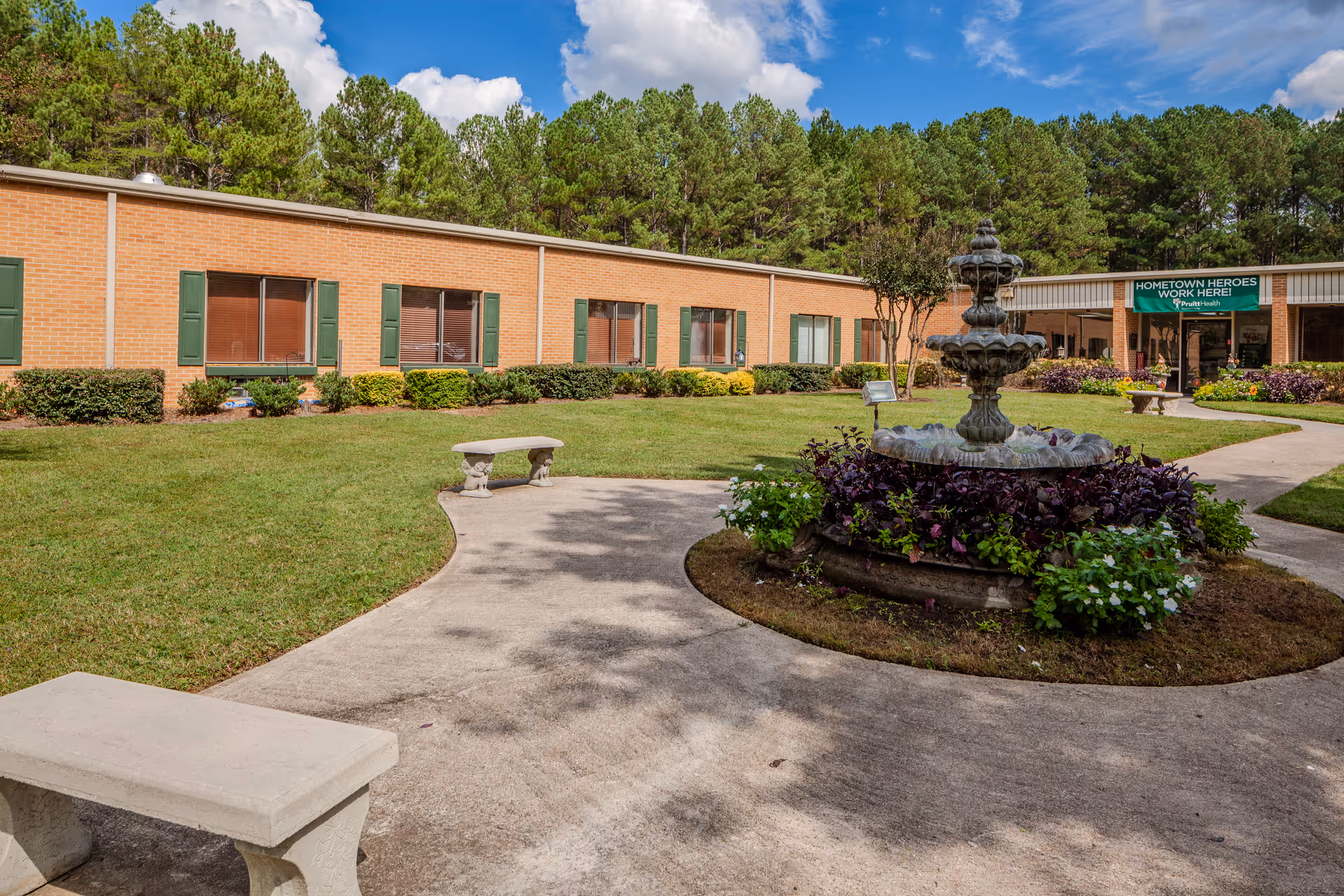 Brick senior living facility courtyard with fountain, benches, lawn, and tree-lined surroundings.