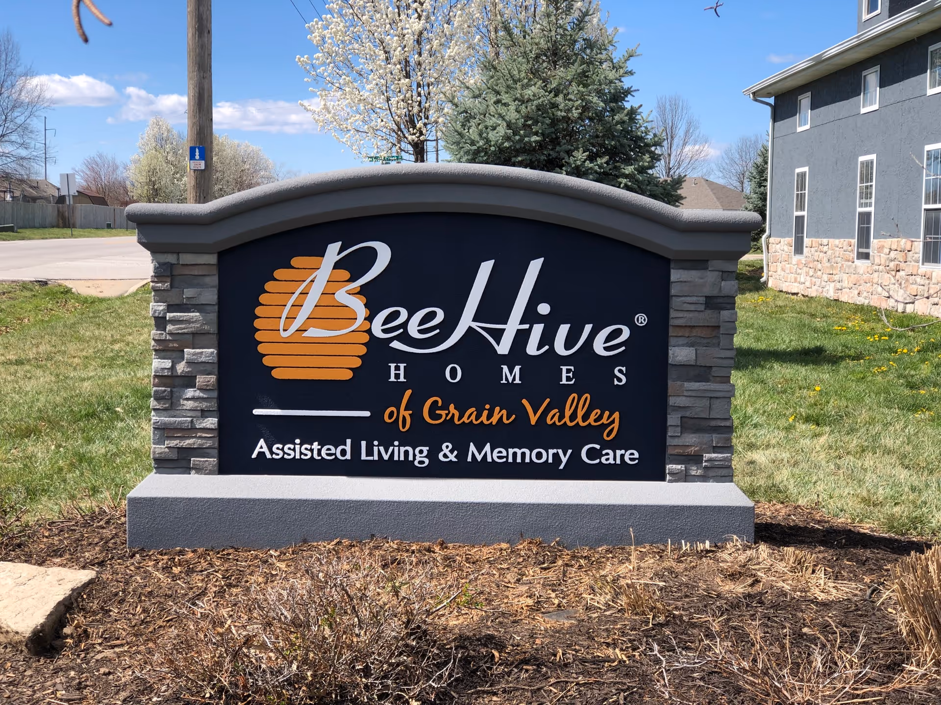 Outdoor stone and concrete sign for BeeHive Homes of Grain Valley, an assisted living and memory care facility, with a building and trees in the background under a blue sky.