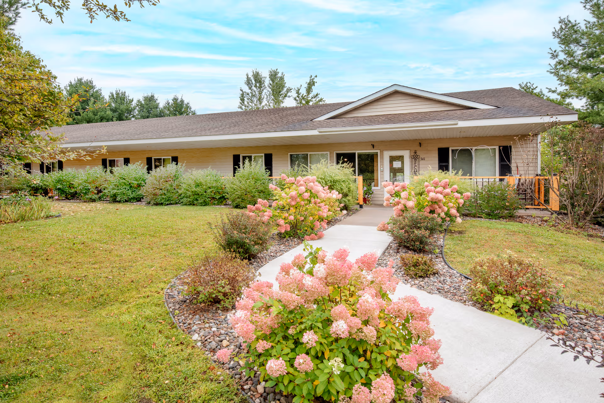 Single-story tan senior living building with a curved walkway and pink flowering shrubs.