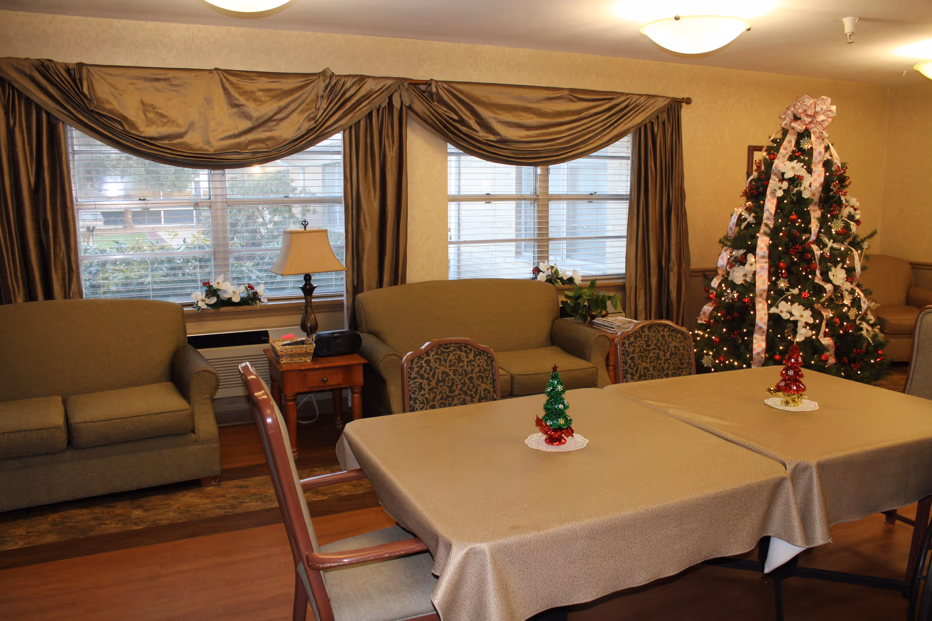 A cozy living room decorated for Christmas with a Christmas tree adorned with lights and ribbons in the corner. The room has two green sofas, a wooden side table with a lamp and some decorations, and a table covered with a beige tablecloth with small Christmas tree decorations on top. Large windows with brown curtains let in natural light.
