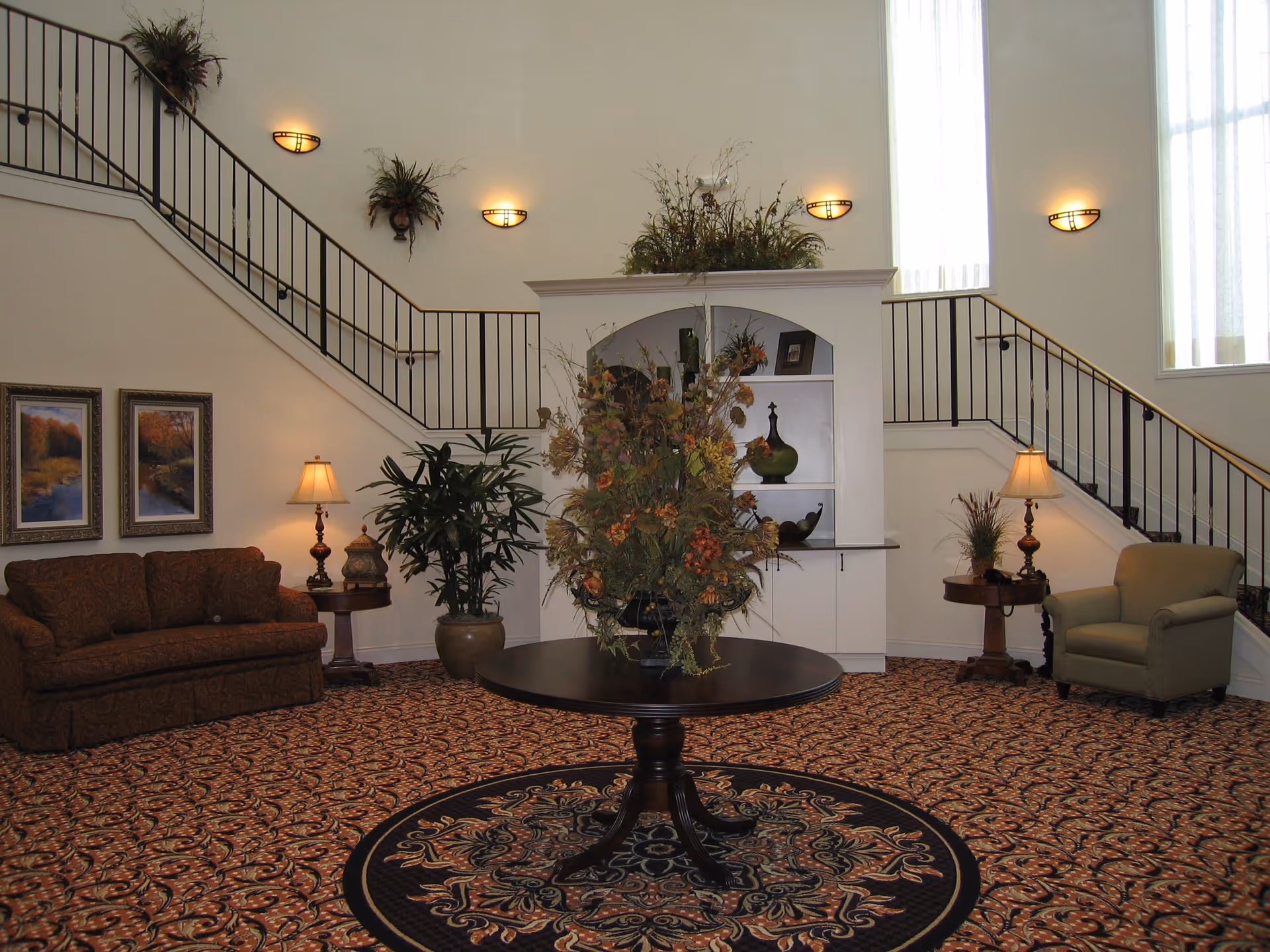 Spacious lobby-style living room with a central round table holding a large floral arrangement, seating areas, patterned carpet, and staircases along the walls.