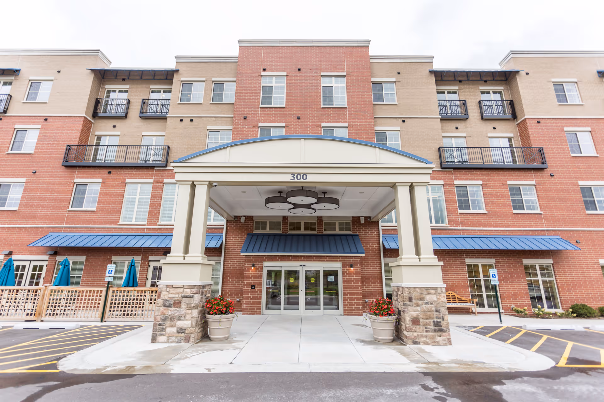 Front exterior view of a multi-story assisted living and memory care facility with a covered entrance supported by columns, large windows, balconies, and a parking area with handicap spaces.