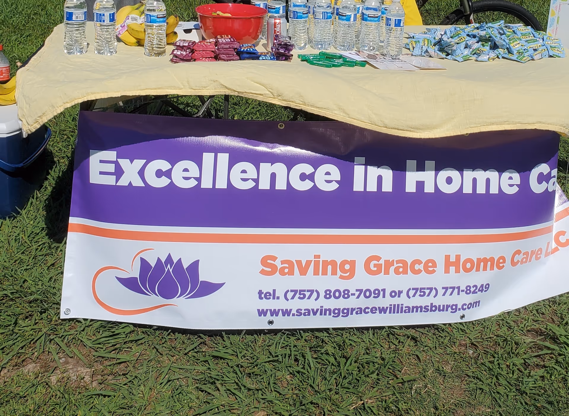 An outdoor table displays water bottles, snacks, and a banner for Saving Grace Home Care LLC.