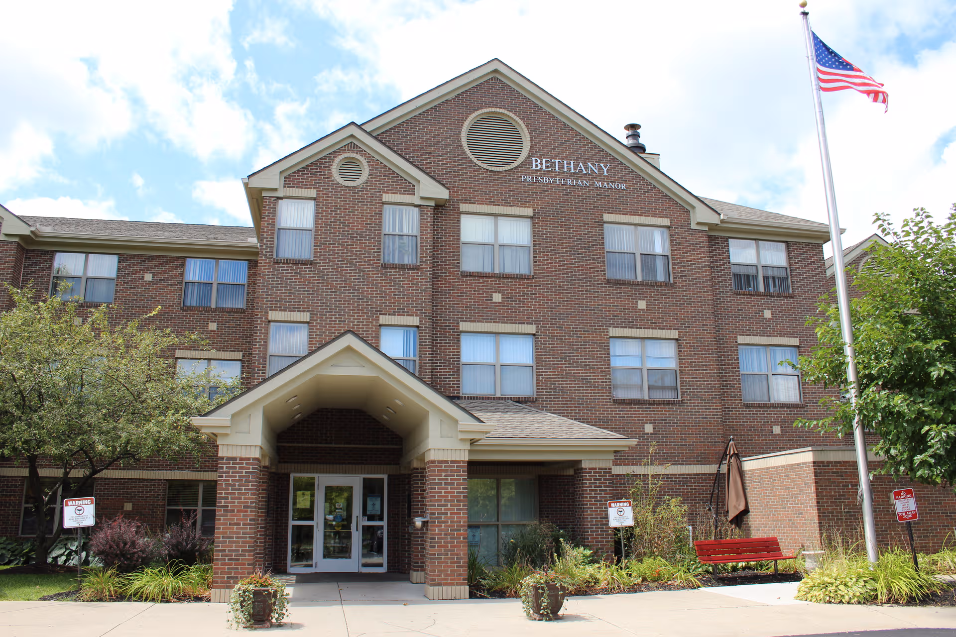 Brick senior living facility facade with central entrance, flag, and surrounding landscaping.