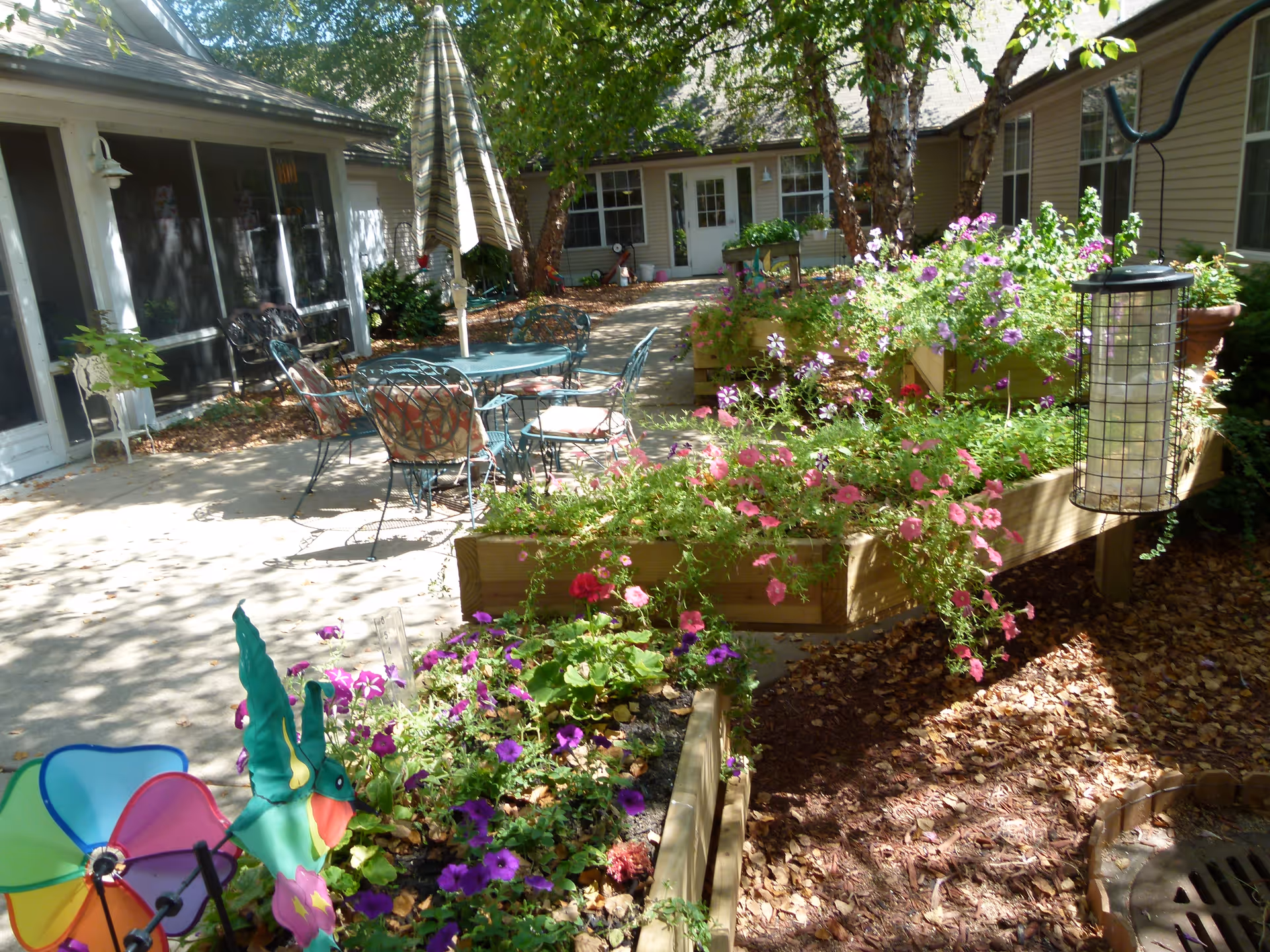 Outdoor patio area with metal chairs and tables, an umbrella, raised garden beds filled with colorful flowers, and a colorful pinwheel decoration. The area is surrounded by buildings with windows and doors, and there are trees providing shade.