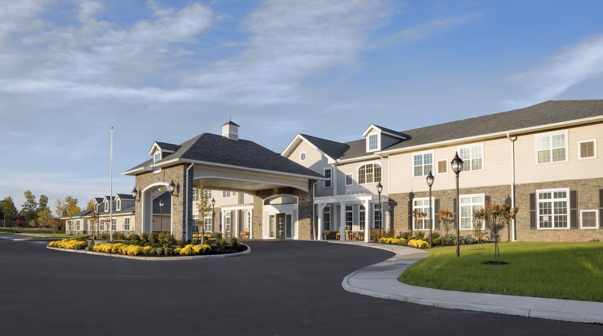 Front entrance of a two-story assisted living facility with a porte-cochère, landscaped driveway, and lawn under a blue sky.