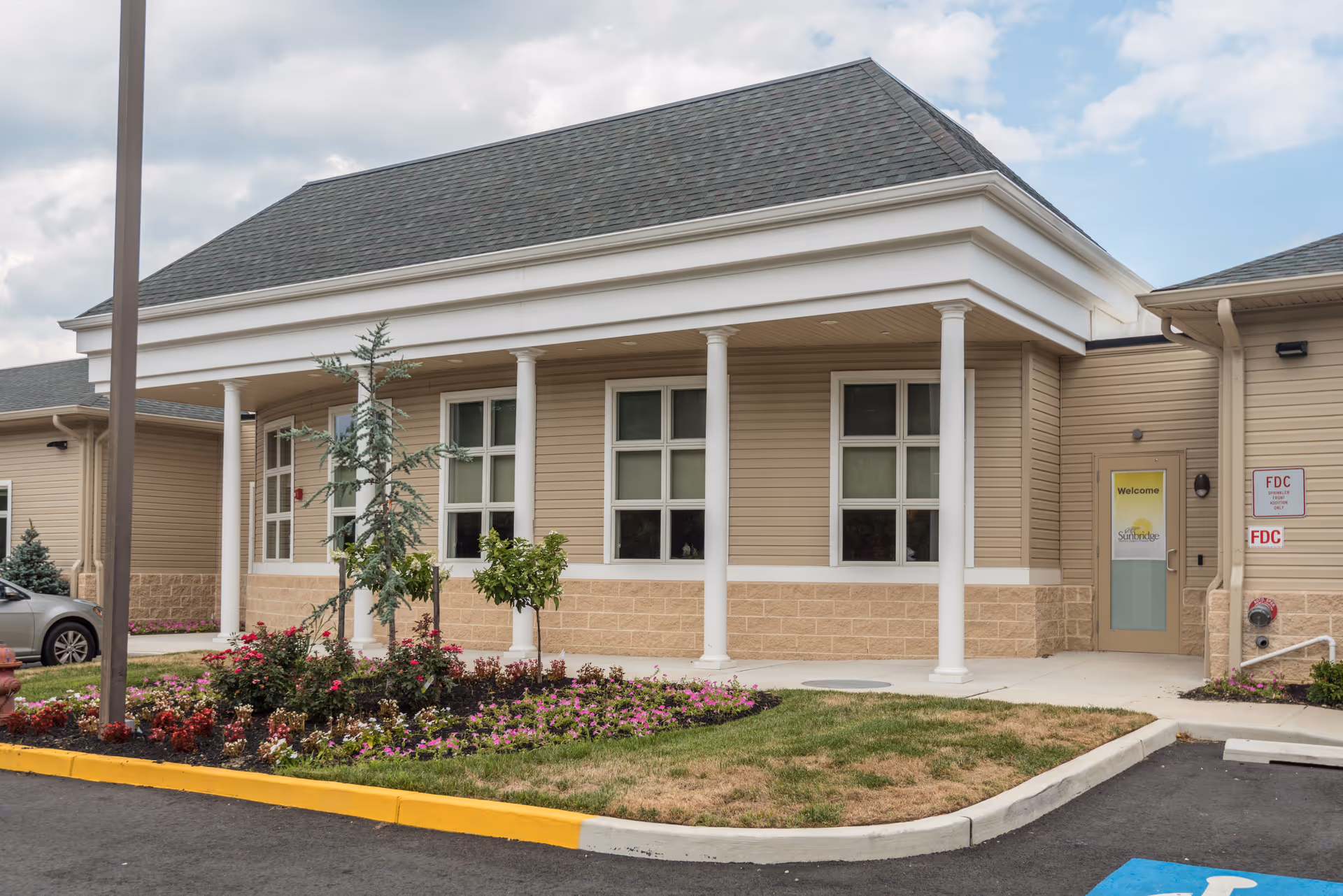 Exterior view of a single-story building with beige siding, white columns, and a gray shingled roof. There is a landscaped flower bed with various plants and flowers in front of the building. A door with a yellow 'Welcome' sign is visible on the right side, along with fire department connection signs. The sky is partly cloudy.
