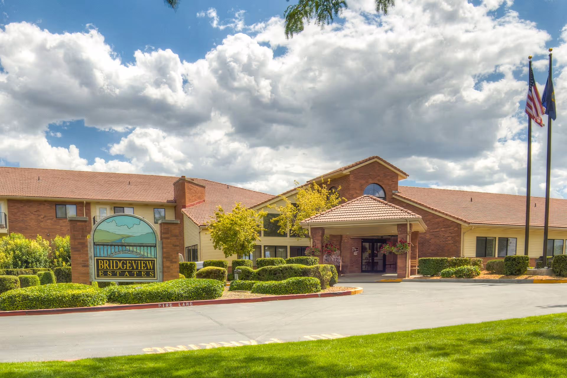 Front exterior of Bridgeview Estates senior living building with a landscaped entrance, sign, and flagpoles under a partly cloudy sky.