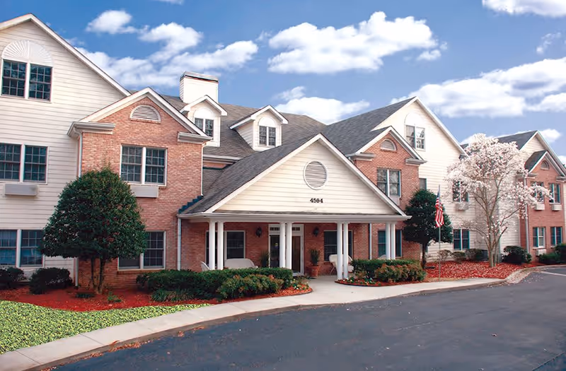 Front exterior of a brick-and-siding senior living building with a covered entrance, landscaped beds, and an American flag.