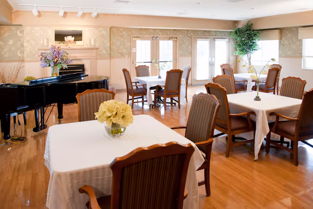 Bright dining room with round tables, white tablecloths, wooden chairs, and a grand piano.