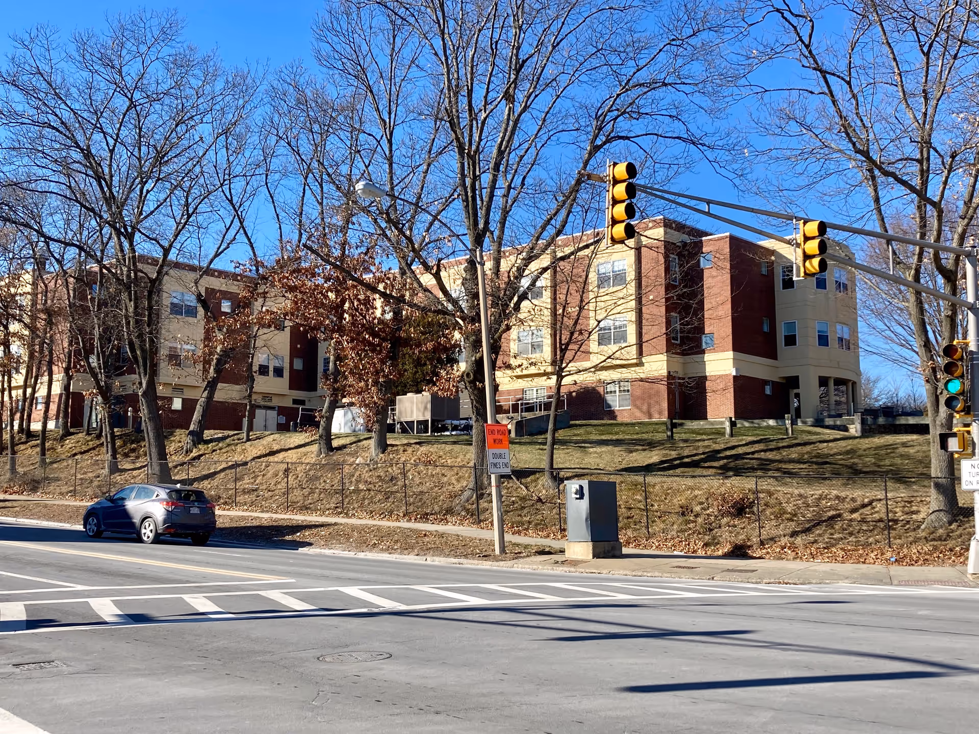 Four-story brick-and-beige senior living building on a grassy hill bordered by a street intersection with traffic lights and a parked car.